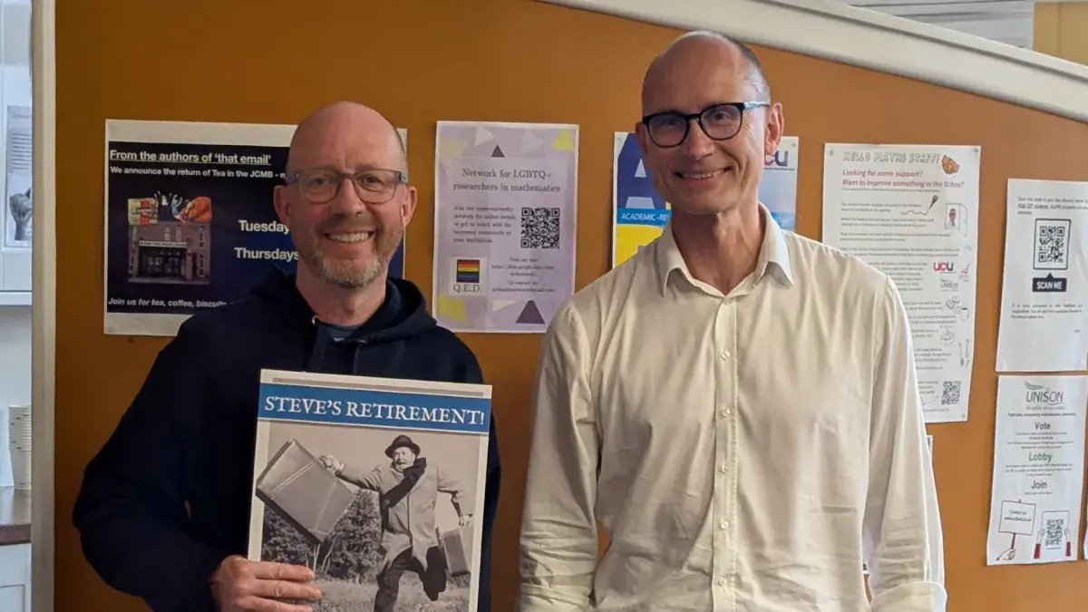 Stev Law and Head of School Bernd Schroers standing side-by-side in the maths common room. Steve is holding a large card, reading "Steve's Retirement!" 