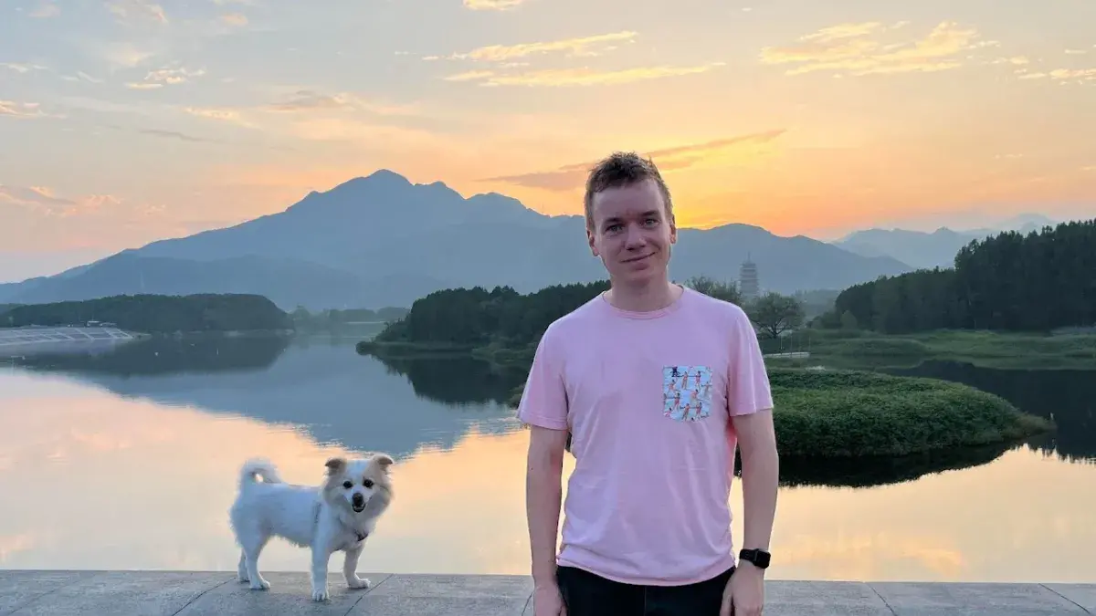 A photo of Ben Davison - he is wearing a pink T-shirt and leaning against a wall. There is a small dog perched on the wall next to him. In the background is a beautiful sunset scene over a lake.