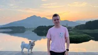 A photo of Ben Davison - he is wearing a pink T-shirt and leaning against a wall. There is a small dog perched on the wall next to him. In the background is a beautiful sunset scene over a lake.