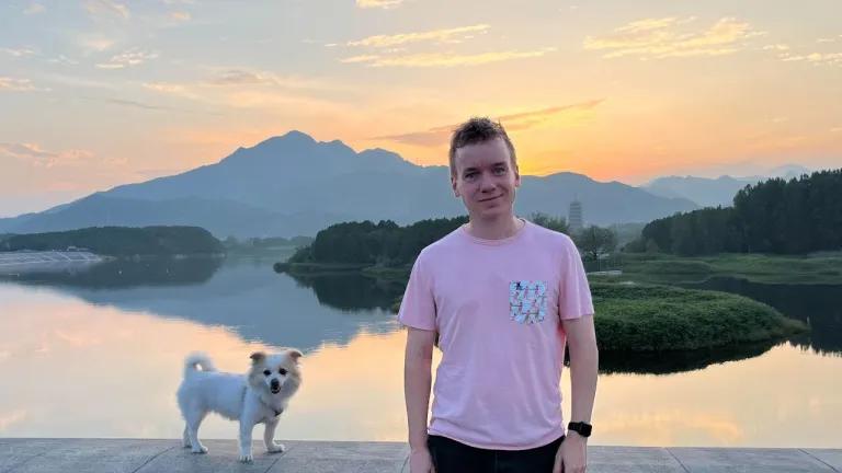 A photo of Ben Davison - he is wearing a pink T-shirt and leaning against a wall. There is a small dog perched on the wall next to him. In the background is a beautiful sunset scene over a lake.