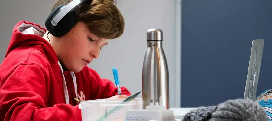 A boy in a red hoodie is doing homework with a laptop and headphones.