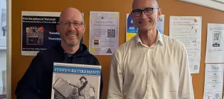 Stev Law and Head of School Bernd Schroers standing side-by-side in the maths common room. Steve is holding a large card, reading "Steve's Retirement!" 