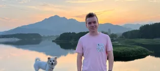 A photo of Ben Davison - he is wearing a pink T-shirt and leaning against a wall. There is a small dog perched on the wall next to him. In the background is a beautiful sunset scene over a lake.