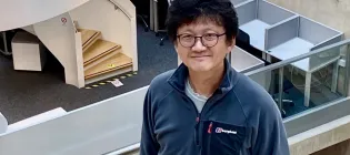 A photo of Minhyong who is looking up at the camera from the stairwell of the Bayes Centre. In the background is a white spiral staircase.