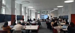 A photograph of the classroom where the hackathon took place. Students are grouped around white desks while a presenter stands in front of a large screen.