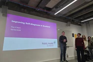 A group of students are standing in front of a large screen presenting their project. The screen reads, "Improving self-diagnosis in Scotland"