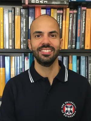 Miguel standing in front of a bookcase wearing a University of Edinburgh-branded polo shirt