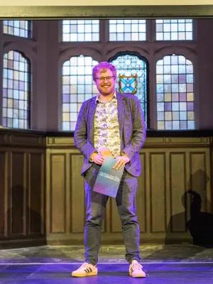 A photo of Jonathan receiving his teaching award. He is standing on a stage in front of a stained glass window in Teviot.