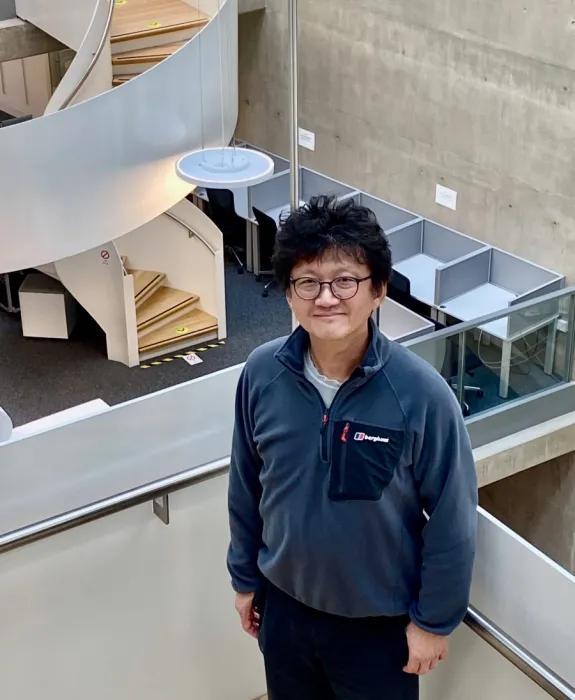 A photo of Minhyong who is looking up at the camera from the stairwell of the Bayes Centre. In the background is a white spiral staircase.