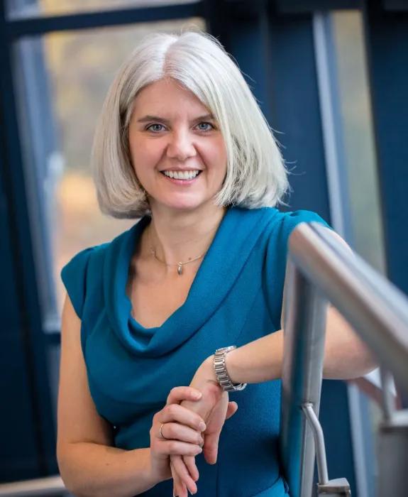 A professional photo of Heather Tewkesbury. She appears to be in a modern stairwell and leaning against a banister while looking at the camera